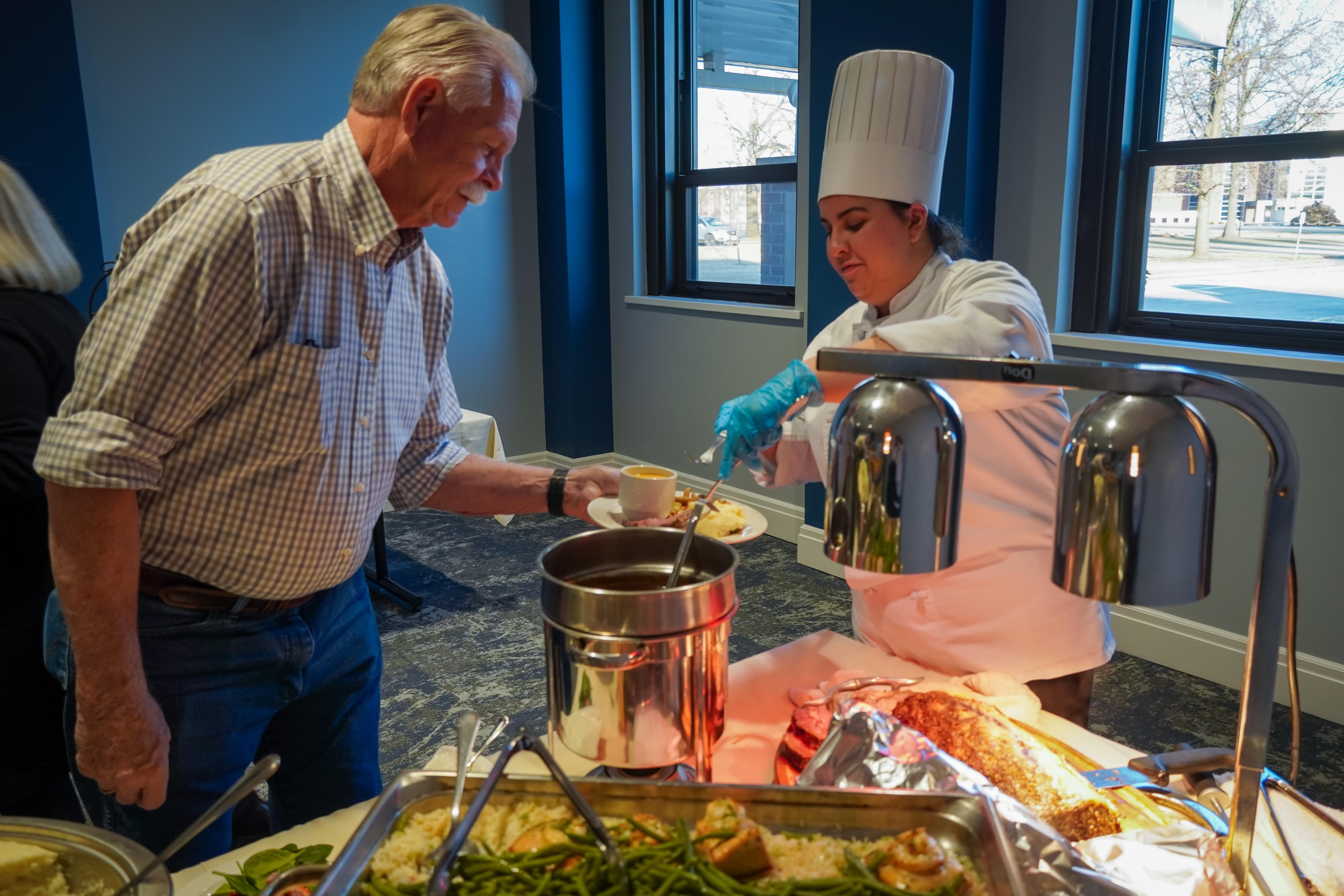 A female student wearing a chef's hat and chef jacket serves an older male diner at a buffet.